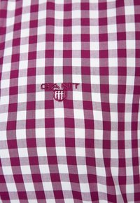 Camicetta GANT con un motivo a scacchi viola e bianco, con un logo ricamato al centro in filo viola coordinato.