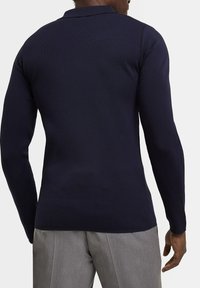 Polo-shirt bleu marine à côtes, à manches longues, avec un col et des fentes sur les côtés. Porté avec un pantalon gris clair, montrant un tissu tricoté texturé.