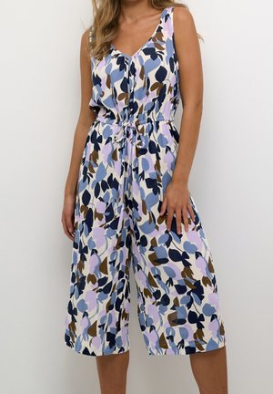 Mouwloze jumpsuit met V-hals, met een patroon van blauwe, paarse en bruine bladeren, een getailleerde taille met bindlint, gedragen door een persoon die voor een vlakke muur staat.