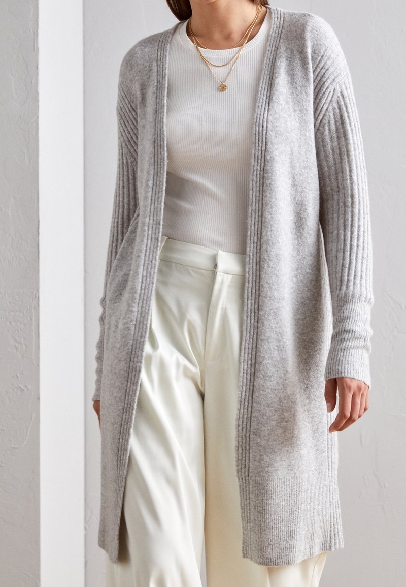 Long cardigan gris clair en maille douce, avec des manches côtelées et un devant ouvert. Porté sur un haut blanc côtelé et un pantalon crème.