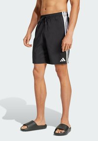 Crne kupaće hlače s bijelim bočnim prugama, elastičnim pojasom s vezicom i malim Adidas logotipom na lijevoj nozi, uparene s crnim natikačama.