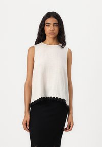Top sans manches en beige clair avec une finition texturée, doté d'un ourlet noir ondulé. Porté avec une jupe moulante noire en côtes.