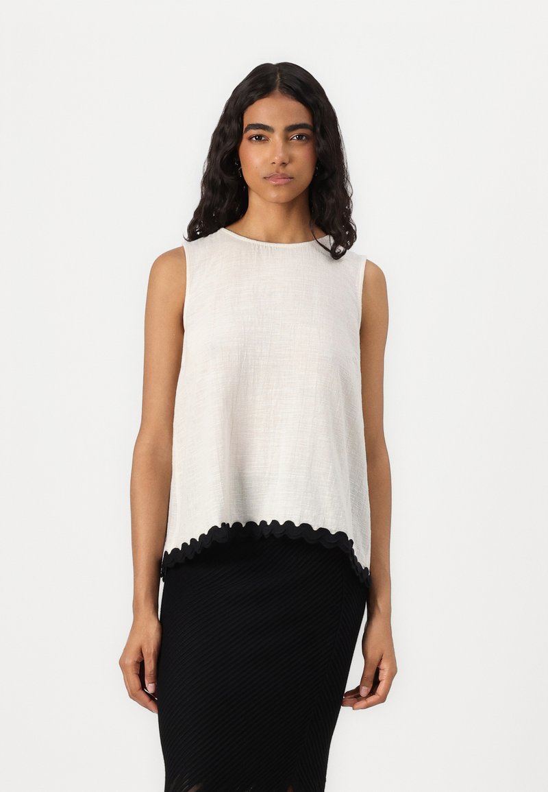 Top sans manches en beige clair avec une finition texturée, doté d'un ourlet noir ondulé. Porté avec une jupe moulante noire en côtes.