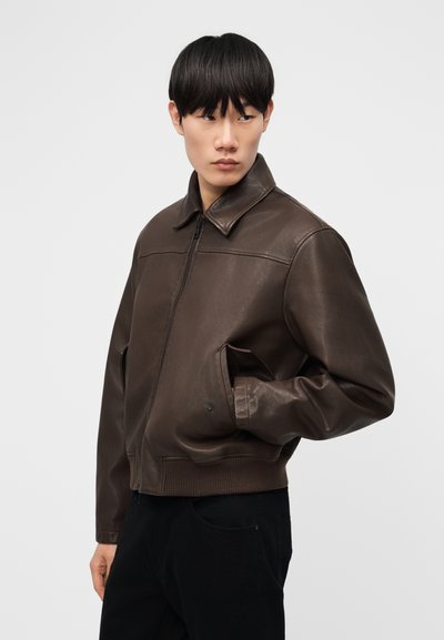 Jeune homme portant une veste en cuir marron foncé avec une fermeture éclair et des poches latérales, regardant vers sa gauche sur un fond blanc uni.