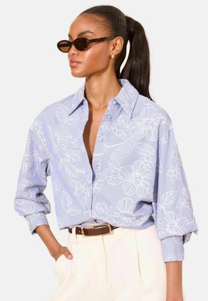 Vrouw met donker haar in een paardenstaart, die schildpadzonnebril draagt, een blauw gebloemd overhemd met knopen, beige broek en een bruine riem.