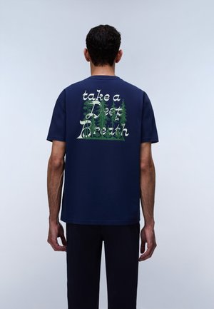 Homme de dos portant un t-shirt bleu marine avec des arbres verts et le texte « take a deep breath » dans le dos sur un fond uni.