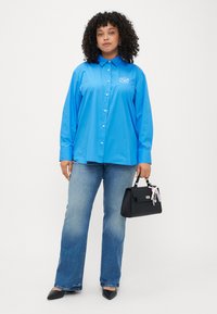 Blå skjorte med lange ermer, kombinert med lyseblå jeans og en svart håndveske med en dekorativ anheng. Svart spiss sko.