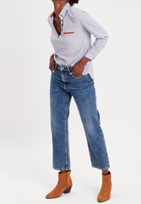 Chemise blanche avec micro-motif bleu, accent orange sur le col, associée à un jean en denim bleu taille haute et des bottines à talons en daim marron.
