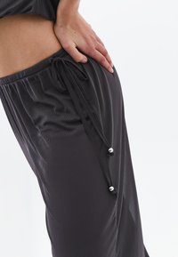 Donkergrijze broek van gladde stof met een tailleband met trekkoord en zilveren metalen accenten. Lichte textuur zichtbaar. Casual fit.