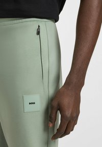 Lichtgroene sportbroek met een ritszak en een vierkante logo-tag met "BOSS." Glad materiaal met een getailleerde pasvorm.