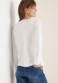 Maglietta bianca a maniche lunghe con scollo rotondo; tessuto morbido e liscio; design a strati con una leggera svasatura all'orlo; abbinata a jeans in denim blu.