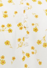 Tissu blanc avec des fleurs jaunes éparpillées au centre rouge, cinq boutons blancs alignés verticalement sur le devant, texture légère.