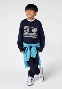 Tengerészkék Champion logós pulóver, derekán megkötött kék pulóver, tengerészkék nadrág és fehér, fekete részletekkel díszített tornacipő. Végig sima textúrájú.