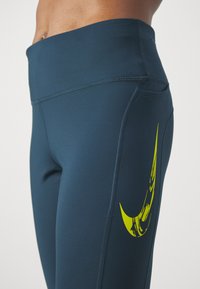 Leggings de sport bleu marine avec une taille haute, présentant un grand logo Nike jaune sur la cuisse gauche, une texture lisse et un tissu extensible.