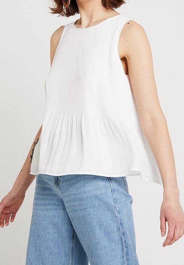 Femme portant un chemisier blanc plissé sans manches et un jean taille haute bleu clair, visible des épaules aux hanches.
