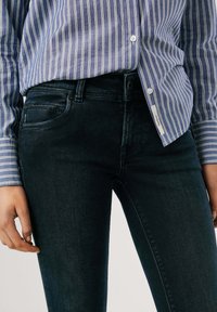 Temno modre skinny jeans z gladko teksturo, kontrastnim šivanjem in sprednjim žepom, nosijo z svetlo modro črtasto srajco.
