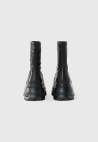 Par de botas de cuero negras hasta el tobillo con suelas gruesas, mostradas desde atrás sobre un fondo gris claro liso.