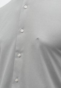 Szürke gombos ing sima textúrával, fehér gombokkal és egy kis hímzett logóval a mellkasnál.