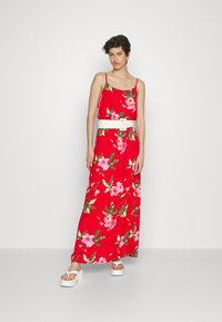 Vestido maxi floral vermelho com alças finas, apresentando padrões de flores rosa e verdes. Acompanhado de um cinto branco e sandálias brancas robustas.