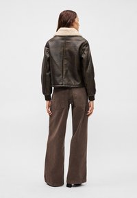 Smeđa kožna bomber jakna s krznatim ovratnikom, uparena sa širokim smešnim hlačama. Jakna ima rebraste manžetne i detalje šivanja.