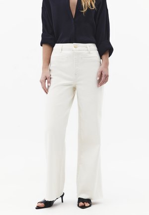 Kvinde iført hvide bukser med højt talje og vidde ben, sort langærmet skjorte med opkørte ærmer og sorte åbne sandaler med hæl.