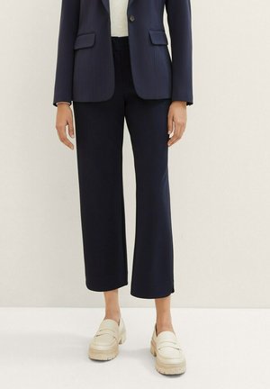 Frau trägt ein dunkelblaues Blazer und eine verkürzte, weit geschnittene Hose, kombiniert mit beigefarbenen, klobigen Plateauslippern.