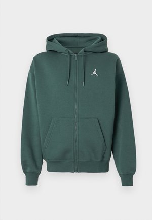 Zelená zapínacia mikina vyrobená z mäkkej látky, má predné vrecko, šnúrky, rebrované manžety a malé biele logo na hrudi.