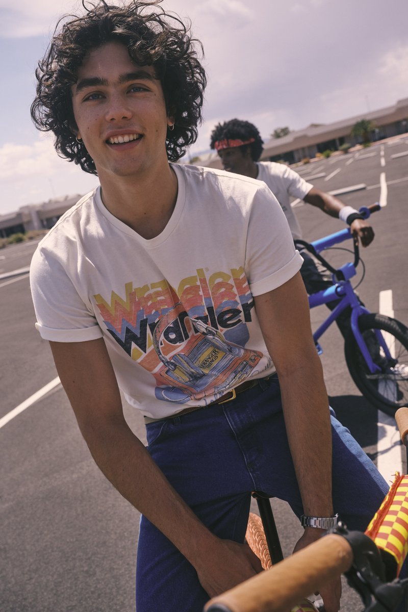 Λευκό μπλουζάκι με πολύχρωμο γραφικό "Wrangler", μπλε τζιν παντελόνι, ασημένιο ρολόι και ποδήλατο με πλεγμένο χερούλι. Υπαίθρια τοποθεσία, καθαρός ουρανός.