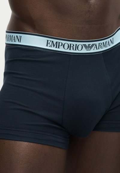 Κοντινό πλάνο ενός ατόμου που φοράει σκούρα μπλε boxer briefs Emporio Armani με λευκή μέση και μαύρο λογότυπο.