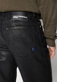 Svarta denimjeans med en slät yta, med rak skärning, bakficka med en blå "K"-emblem och läderlogga märkt "KARL LAGERFELD JEANS."