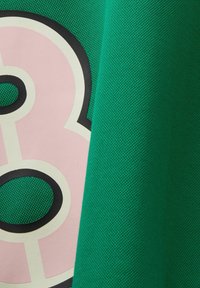 Grön texturerad tyg hängande över en rosa och vit grafik med en stor svart kontur av en bokstav eller siffra.