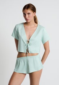 Mintgroen loungewear-set met een cropped top met een diepe V-hals en striksluiting, gecombineerd met bijpassende shorts. Gladde satijnen textuur.