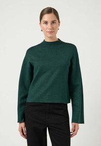 Ciemnozielony, strukturalny sweter z wysokim kołnierzem i szerokimi rękawami, noszony na czarnych spodniach. Prezentuje luźny krój i równą linię dołu.