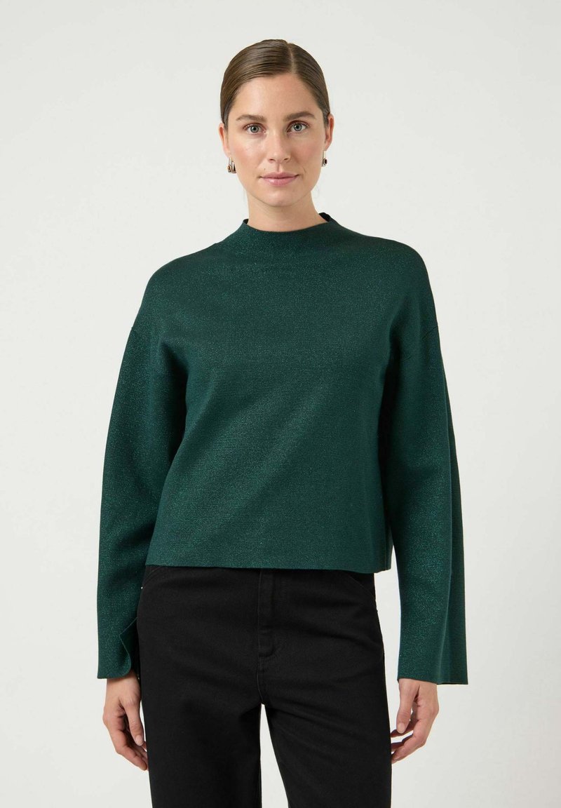 Ciemnozielony, strukturalny sweter z wysokim kołnierzem i szerokimi rękawami, noszony na czarnych spodniach. Prezentuje luźny krój i równą linię dołu.