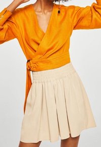 Femme portant un chemisier cache-cœur orange vif à manches longues et une jupe plissée taille haute beige, posant avec les bras levés.