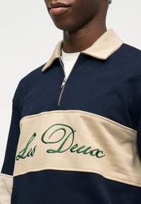 Άνδρας που φοράει μπλε ναυτικό και μπεζ φούτερ με φερμουάρ και γιακά, με πράσινο κεντητό κείμενο «Les Deux» στο στήθος.