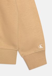 Manga de sudadera de algodón beige con puño acanalado. Presenta el logo bordado "cK" en blanco. Textura suave, diseño minimalista.