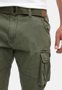 Homme portant un pantalon cargo vert olive avec une ceinture en tissu assortie, un t-shirt blanc rentré et la main détendue le long du corps.