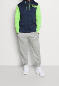Navy and neon green zip-up hoodie with a Seahawks logo on the chest, paired with gray sweatpants and white sneakers.