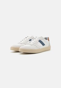 Zapatillas deportivas blancas con una suela beige, que presentan una tela texturizada y cuero liso. Acentos azules a lo largo de los lados y una pestaña del talón marrón.