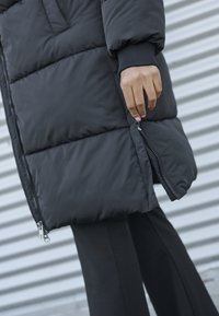 Fekete puffert jacket, cipzárral záródik, és steppelt mintázattal rendelkezik. Bordás mandzsettákkal és oldalsó cipzárral készült. A modellen a kezét a kabát mellett helyezte el.
