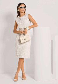 Weißes, strukturiertes Kleid mit einer figurbetonten Silhouette, ergänzt mit einer kleinen cremefarbenen Handtasche, einem breiten Armreif und weißen Stilettosandalen.