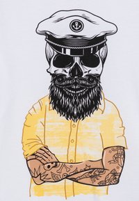 Diseño gráfico de un cráneo con barba y bigote, usando un gorro de capitán, sobre una camiseta amarilla con tatuajes en los brazos, sobre un fondo blanco.