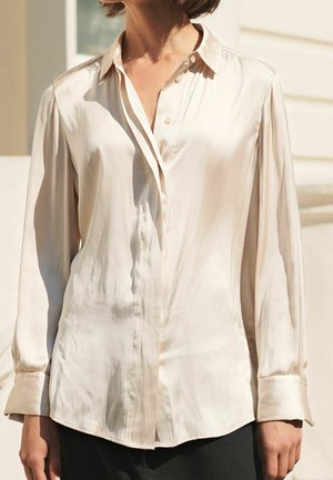 Femme portant une chemise boutonnée à manches longues en satin ivoire avec col et poignets, se tenant devant un fond neutre baigné de lumière naturelle.