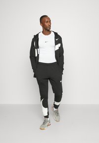 Sudadera negra de Nike que consiste en una chaqueta con capucha y acentos reflectantes, una camiseta blanca ajustada y pantalones negros con detalles blancos.