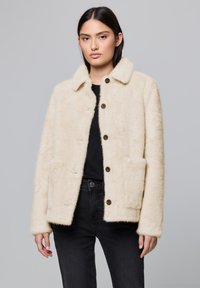 Veste en fausse fourrure crème avec un col, dotée de deux poches avant et de boutons ronds marron. Portée sur un haut noir et un jean.