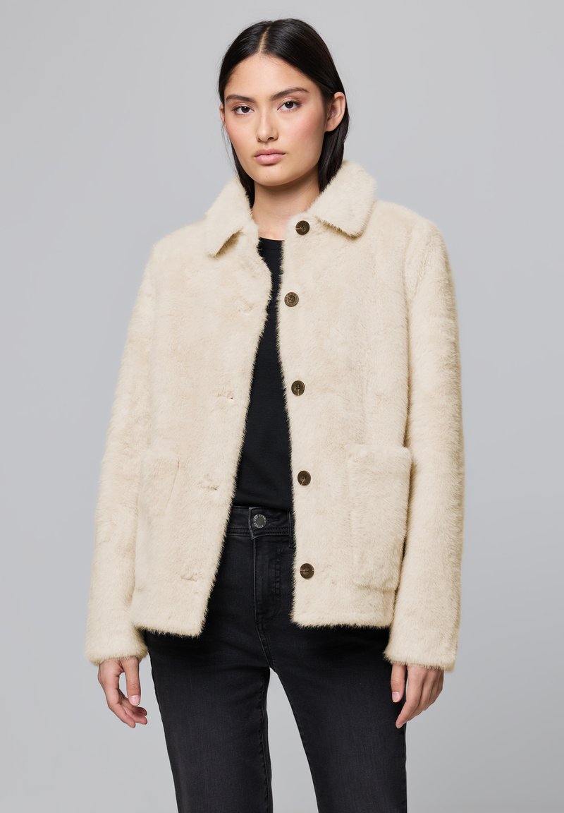 Veste en fausse fourrure crème avec un col, dotée de deux poches avant et de boutons ronds marron. Portée sur un haut noir et un jean.