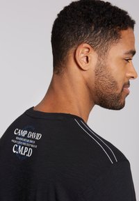 Svart t-shirt med vit accentstickning och varumärkeslogotyp på baksidan. Materialet verkar lätt och texturerat, lämpligt för avslappnad klädsel.