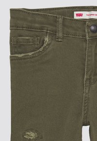 Los vaqueros super ajustados Levi's 710 en verde oliva presentan una textura de mezclilla, una costura de bolsillo deshilachada y un cierre de botón con la etiqueta de la marca visible.