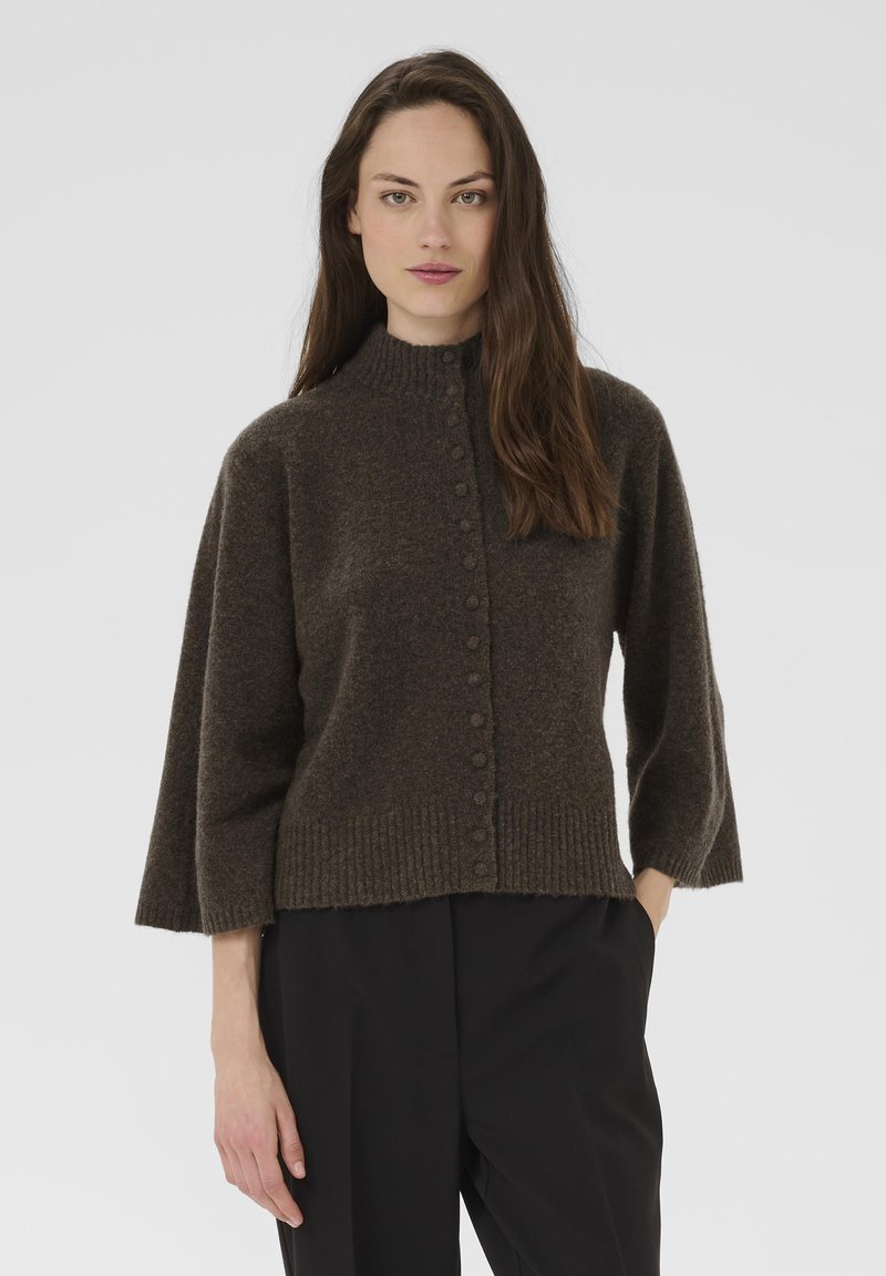 Hosszú barna hajú nő sötétbarna, gombos gyapjúpulóverben, széles ujjakkal és fekete nadrágban, egy sima háttér előtt áll.